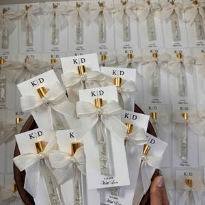 Può includere: Una collezione di fiale in vetro trasparente con tappi dorati, fissate su cartoncini bianchi con le lettere KD, il testo Bridal Shower, la data 9.19.2026 e la frase With Love. Ogni fiala è decorata con un fiocco di nastro bianco e una perla.