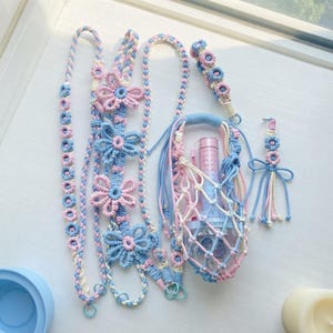 May include: A collection of handmade macrame accessories in pastel pink, blue, and white, including a long braided strap with flower motifs, a net-style bottle holder containing a light stick, a small wristlet, and a decorative tassel charm.