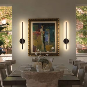 May include: Two vertical wall sconces with a matte black finish and integrated LED light strips. Each fixture features a central circular mounting plate with a long, slender rod extending above and below, emitting a warm glow against the wall.
