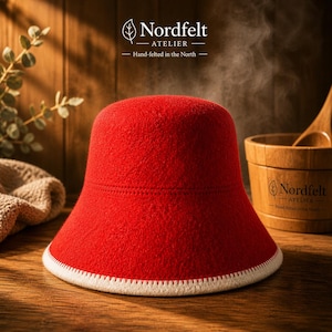 Könnte beinhalten: Ein leuchtend roter Filzhut mit weißem, genähtem Rand. Der Hut ist im Bucket-Stil gehalten, mit dem Text "Nordfelt ATELIER - Hand-felted in the North" sichtbar. Der Hut steht auf einer Holzoberfläche.