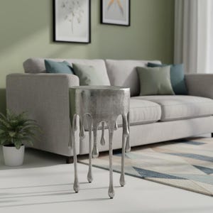 Puede incluir: Una mesa auxiliar plateada con un diseño único. La mesa tiene una parte superior cilíndrica y presenta un efecto de goteo en los lados, con patas alargadas en forma de lágrima. La mesa está en un salón.