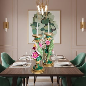 May include: Two ornate, emerald green and gold candle holders with parrot and floral designs. The candle holders are placed on a dark wood dining table set for a meal, with green velvet chairs. A gold chandelier hangs above.