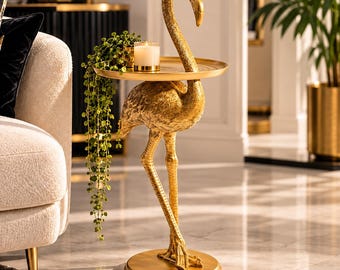 May include: A gold-colored flamingo-shaped side table with a circular tabletop. The flamingo stands on a round base, and the tabletop features a textured surface. A small white candle in a glass holder and a trailing green plant sit on the table.