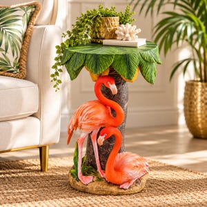May include: A round side table featuring a palm tree trunk base with two bright pink flamingos. The tabletop is designed to look like large green palm leaves. The base is textured like bark and sits on a sandy-colored circular foundation.