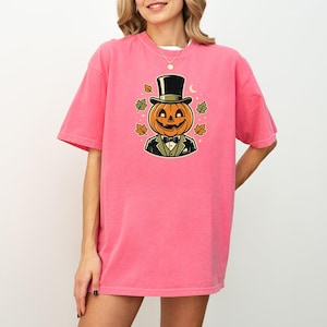 May include: A bright pink short-sleeve t-shirt featuring a central graphic of a jack-o'-lantern wearing a black top hat and a tuxedo with a bow tie. The pumpkin character is surrounded by small colorful autumn leaves and tiny white stars.