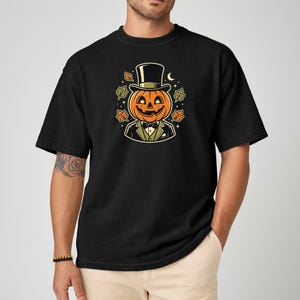 May include: A black short-sleeve t-shirt featuring a graphic of a smiling jack-o'-lantern wearing a black top hat and a formal suit with a bow tie. The pumpkin is surrounded by small autumn leaves, stars, and a crescent moon illustration.