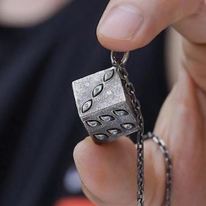 Op de afbeelding: Een zilveren hanger in de vorm van een dobbelsteen, versierd met oogvormige ontwerpen met blauwe details. De hanger hangt aan een ketting. De dobbelsteen wordt tussen vingers gehouden, tegen een donkere achtergrond.
