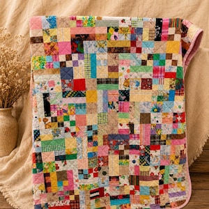 May include: A rectangular patchwork quilt featuring a dense grid of multi-colored fabric squares and rectangles. The textile displays a variety of floral, plaid, and solid patterns in shades of pink, blue, yellow, green, and red, finished with a pink border.