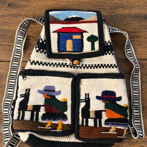 May include: A woven backpack with a cream-colored body and black and white striped straps. The bag features three pockets with colorful embroidered scenes of houses, mountains, and figures with llamas. The bag has a button closure.