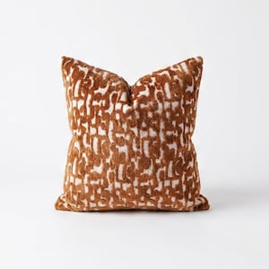 Op de afbeelding: Vierkant sierkussen met een bruin en wit abstract patroon. Het kussen heeft een zacht, getextureerd uiterlijk en is waarschijnlijk gemaakt van een pluche stof. Het ontwerp heeft een herhalend patroon van onregelmatige vormen.