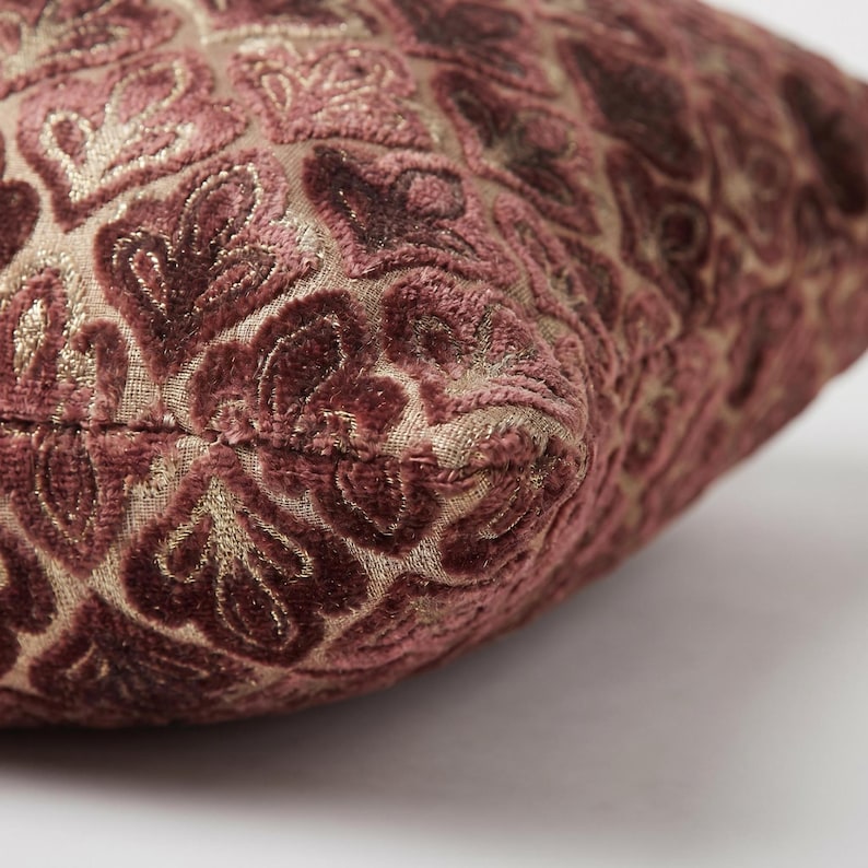 May include: A square throw pillow featuring a raised, velvet-like floral pattern in shades of deep mauve and burgundy. The fabric base is a light tan, woven material that contrasts with the soft, textured, and plush velvet floral motifs.