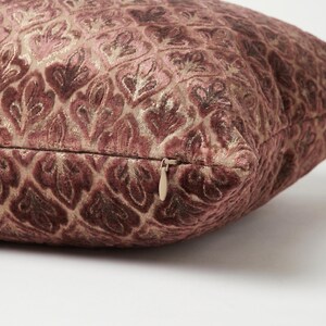 May include: A square throw pillow featuring a textured, raised velvet pattern in shades of dusty rose and mauve against a metallic gold background. The pillow includes a concealed zipper closure with a small, matching pull tab on the side edge.