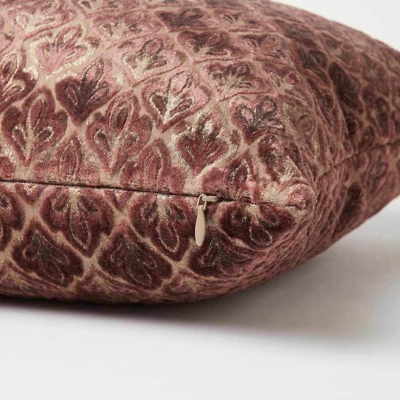 May include: A square throw pillow featuring a textured, raised velvet pattern in shades of dusty rose and mauve against a metallic gold background. The pillow includes a concealed zipper closure with a small, matching pull tab on the side edge.