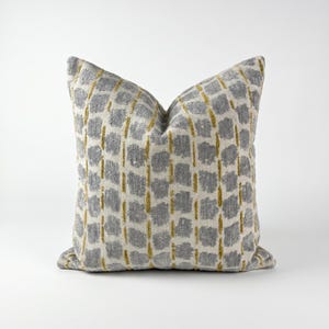 May include: A square throw cushion featuring a textured pattern of grey rectangular shapes arranged in vertical columns, separated by thin, vertical lines of mustard yellow thread against a light cream-coloured fabric background.