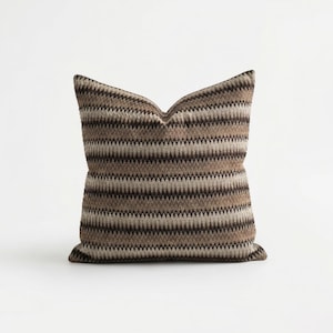 Puede incluir: Un cojín cuadrado decorativo con un patrón de zigzag marrón y beige. El cojín tiene un aspecto suave y texturizado y está diseñado para la decoración del hogar. La paleta de colores incluye tonos de marrón, beige y crema.