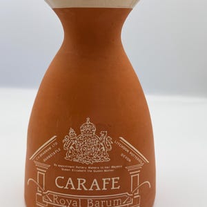 May include: A terracotta carafe with a flared rim and a cream-colored interior. The front features a white printed royal crest, the text CH BRANNAM LTD BARNSTAPLE, LITCHDON POTTERIES DEVON, CARAFE, Royal Barum Ware, and By appointment Pottery Makers.