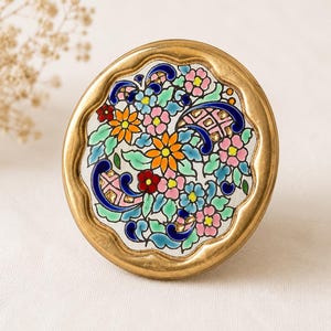 Könnte beinhalten: Ein runder, dekorativer Möbelknopf mit einem goldfarbenen, gewellten Rand. Die Mitte zeigt ein farbenfrohes Blumenmuster mit orangefarbenen, rosa, blauen und grünen Blumen. Der Knopf ist wahrscheinlich für einen Schrank oder eine Schublade.