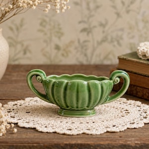 May include: A green ceramic bowl with two ornate handles, resting on a cream-colored doily. The bowl has a scalloped rim and vertical ridges. A vase with dried flowers and a stack of books are in the background.
