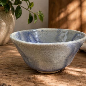 May include: A handcrafted ceramic bowl with a light blue and grey marbled glaze. The bowl has a wide, shallow shape and is placed on a wooden surface. The bowl is likely used for serving food or as a decorative item.