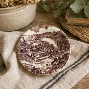 May include: A round ceramic saucer featuring a brown and white transferware print of a rural village scene. The illustration depicts stone buildings, rolling hills, and two figures on horseback accompanied by two small dogs on a country road.