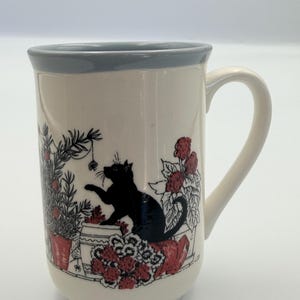 May include: A cream-coloured ceramic mug with a light blue rim and a handle. The exterior features a black silhouette illustration of a cat playing with a hanging ornament amidst potted plants with red flowers and green foliage.
