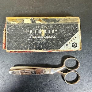 Könnte beinhalten: Eine Zickzackschere aus silberfarbenem Metall mit zwei Fingerringen und einer zentralen Schraube. Die Klinge ist mit dem Text PINKING SHEARS CORP N.Y. graviert. Die Schere wird von einer rechteckigen schwarzen Schachtel mit der Aufschrift PINKIE Pinking Shears begleitet.