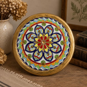 May include: A circular decorative plate with a gold-coloured rim and a vibrant floral design. The pattern includes red, yellow, blue, green, and white details. The plate is likely ceramic or porcelain, adding a touch of elegance to any room.