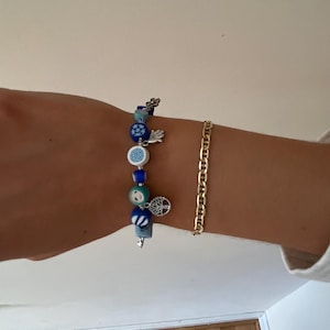 Op de afbeelding: Twee armbanden om een pols. Eén is een kralenarmband met blauwe en witte kralen, bedels en een zilveren levensboom bedel. De andere is een gouden kettingarmband.