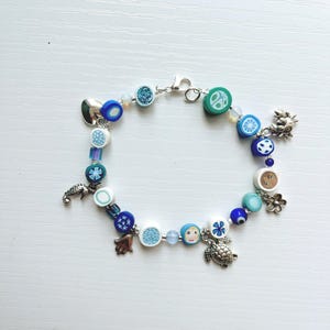 Puede incluir: Una pulsera de dijes con una variedad de cuentas coloridas en tonos azules, blancos y verdes. La pulsera incluye dijes de metal plateado con formas de caballito de mar, tortuga, flor, mano y cangrejo, asegurada con un cierre de mosquetón.