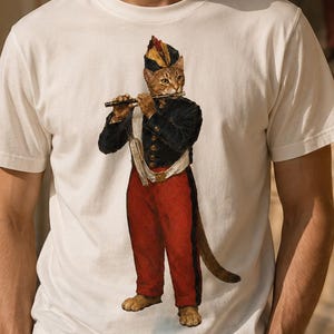 Può includere: Una t-shirt bianca a maniche corte con girocollo caratterizzata da una stampa grafica di un gatto vestito con una giacca in stile militare blu scuro, pantaloni rossi e un cappello abbinato, mentre suona il flauto. Il gatto è raffigurato in piedi.