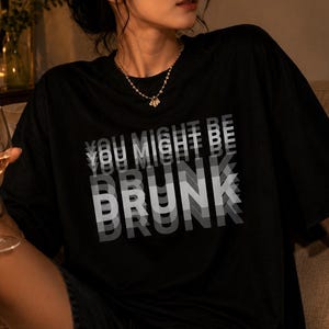 Peut inclure: Un t-shirt noir à manches courtes avec le texte blanc "YOU MIGHT BE DRUNK" imprimé sur la poitrine. Le lettrage présente un effet de flou de mouvement superposé, créant un motif visuel répétitif et décalé qui semble varier en profondeur.