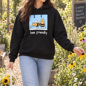 Könnte beinhalten: Ein schwarzer Kapuzenpullover mit einem Grafikdruck von zwei gelb-schwarzen Bienen auf hellblauem Hintergrund und dem Schriftzug "bee friendly" darunter. Der Hoodie verfügt über eine Kängurutasche auf der Vorderseite und Kapuzenkordeln.
