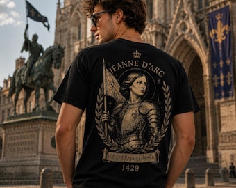 Può includere: Una t-shirt nera a maniche corte con una grafica color oro sul retro. La stampa raffigura Giovanna d'Arco in armatura che regge una bandiera, circondata da una corona d'alloro, con il testo Jeanne d'Arc, Foi Courage France e l'anno 1429.