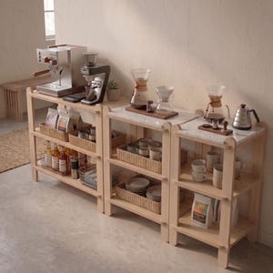 May include: A light wood shelving unit with three tiers, displaying coffee-making equipment and supplies. The top shelf holds a silver espresso machine and grinder. The lower shelves feature baskets with coffee beans, bottles, and cups. The unit has a marble countertop.