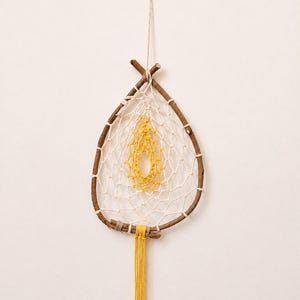 Pode incluir: Um adorno de parede em forma de gota construído com uma moldura de galho de madeira castanha. O interior apresenta um padrão de rede tecido em branco com um centro de croché amarelo. Uma borla de fio amarelo pende da base da moldura.