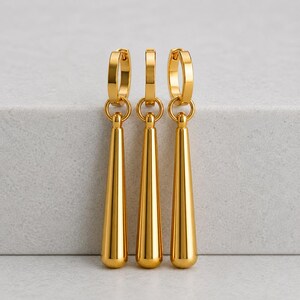 Op de afbeelding: Drie goudkleurige, kegelvormige oorhangers. Elke oorbel heeft een cirkelvormige hoepel aan de bovenkant en een lange, gladde, druppelvormige hanger. De oorbellen worden gepresenteerd tegen een neutrale achtergrond.