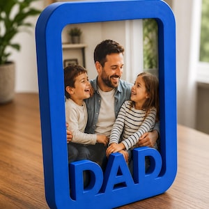 Może przedstawiać: Prostokątna niebieska ramka na zdjęcia ze słowem DAD wkomponowanym w dolną krawędź. Ramka zawiera fotografię uśmiechniętego dorosłego z dwójką dzieci, chłopcem i dziewczynką, którzy śmieją się razem w jasnym, domowym wnętrzu.