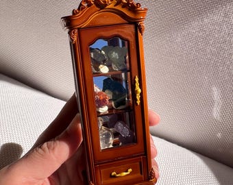Puede incluir: Una vitrina miniatura de color marrón con detalles tallados ornamentados, una puerta de cristal y un pequeño cajón inferior. El mueble cuenta con herrajes dorados y contiene varios especímenes minerales pequeños y variados en sus estantes.