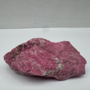 May include: A raw, unpolished mineral specimen featuring a vibrant magenta and deep pink coloration with visible white and grey crystalline inclusions throughout its rough, uneven, and textured surface, showcasing natural geological formations.