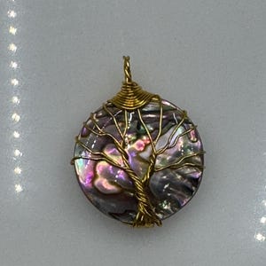 May include: A circular pendant featuring an iridescent abalone shell disc wrapped in gold-colored wire. The wire is intricately twisted to form the shape of a tree with branches extending across the surface of the shell, topped with a wire loop.