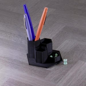 May include: A black desk organizer with multiple compartments, holding a purple and an orange pen. A small tray contains green push pins. The organizer sits on a light gray wooden surface.