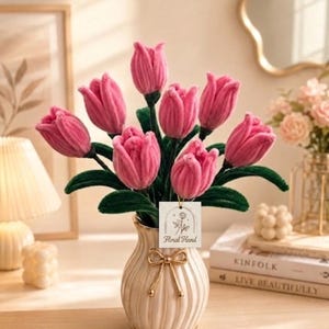 May include: A bouquet of pink tulip flowers in a cream-colored vase with a gold bow. The vase sits on a wooden surface. A tag that says "Floral Hand" hangs from the flowers. The tulips have green stems and leaves.