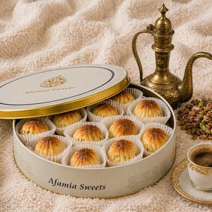 Könnte beinhalten: Eine ovale Dose mit "Afamia Sweets", gefüllt mit Gebäck in Papierförmchen. Eine goldgeränderte Kaffeetasse mit Untertasse, eine Messing-Kaffeekanne und Pistazien sind ebenfalls zu sehen. Der Deckel der Dose ist geöffnet.