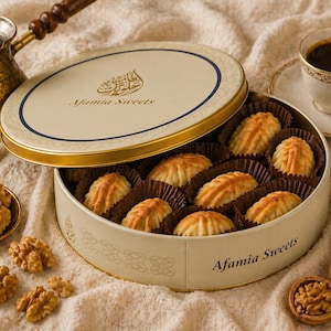 Op de afbeelding: Een open, ronde doos Afamia Sweets, gevuld met goudbruine gebakjes, staat naast een kopje koffie en walnoten. De doos heeft een crèmekleurig deksel met een blauwe rand en gouden letters. Op de achtergrond staat een koperen koffiepot.