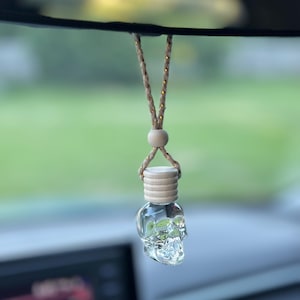 May include: Clear glass skull-shaped car air freshener hanging from a brown and gold rope. The air freshener has a wooden cap and a small wooden bead. The background is blurred, showing a green outdoor scene.