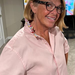 May include: A light pink button-down shirt with a patterned collar. The collar features a colorful floral design. The shirt has short sleeves and is buttoned up the front. The person is wearing glasses with a black and tortoise shell pattern.
