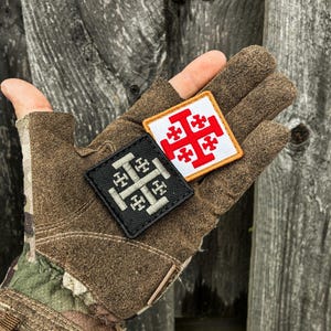 May include: Two square embroidered patches featuring a Jerusalem cross design. One patch has a white background with a red cross and an orange border, while the other patch has a black background with a light gray cross and a black border.