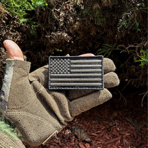 May include: A rectangular patch featuring the American flag in shades of gray and black. The patch is held in a brown glove, with a background of green foliage and red mulch.