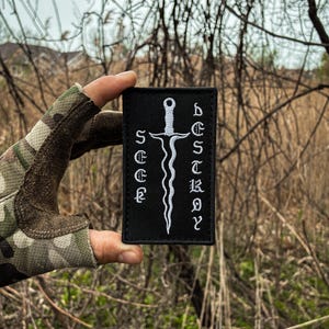 May include: Black rectangular patch with white embroidered text and a stylized sword design. The patch reads "DESTROY" vertically and "SEE" horizontally. The patch is held up against a blurred outdoor background.
