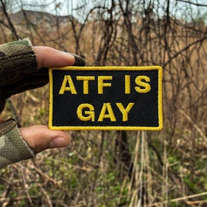 May include: A rectangular embroidered patch with a black background and a yellow border. The text "ATF IS GAY" is stitched in yellow capital letters across the center of the patch. The patch features a textured fabric surface and clean edges.